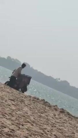 A medium, low-angle shot shows a man with dark hair on a sandy beach next to the ocean. He is wearing a light-colored shirt and dark pants, and is positioned next to a motorcycle that is carrying a large container. The motorcycle is on the sand, facing the water. The man appears to be tending to the load on the motorcycle. The water is a sparkling turquoise color. In the background, across the water, there is land covered in trees. The sky is a hazy white.