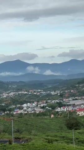 The video pans across a lush, hilly landscape dotted with houses. In the background, mountains fade into the horizon under a cloudy sky. The houses, mostly white with red or blue roofs, are nestled among the greenery. The vegetation appears to be a mix of trees and what might be cultivated fields. The overall impression is a peaceful, rural scene.