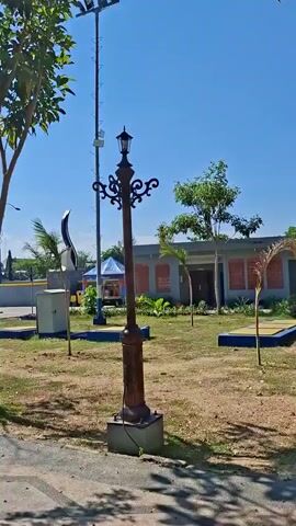 The video begins with a close-up of a decorative lamppost with ornate scrollwork and a glass lantern, standing on a concrete base. The lamppost is brown and weathered, suggesting an older design. In the background, a tall modern lamppost with multiple floodlights is visible. The scene is outdoors, with a clear blue sky above.

The camera then pans to the left, revealing a park-like setting. Several young trees with sparse green leaves are scattered across a dry, grassy area. In the distance, there are some structures including a building with a grey roof and a blue trim, and a large monument with two tall, light-colored pillars. There are also some black, curved sculptures or decorations scattered throughout the park. A grey utility box is visible near one of the sculptures.

The pan continues, showing more of the park, including benches and the walkway. The light suggests it is daytime, likely morning or afternoon. There are no people visible in the video. The overall impression is of an outdoor public space, possibly a plaza or park, with a mix of traditional and modern elements in its landscaping and lighting.
