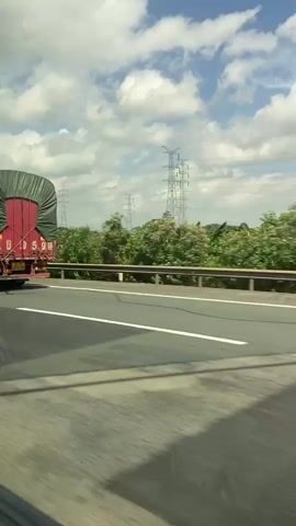 The video shows a view from a vehicle traveling on a highway. A large red and green truck is visible in the foreground, traveling in the same direction. The truck has a green tarp covering its cargo and a license plate that reads "KB 059X". In the background, there are trees, power lines, and a blue sky with white clouds. The perspective suggests the video was filmed from a passenger seat or by someone looking out a side window.