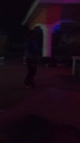 The video shows the inside of a dimly lit outdoor bar at night. There are a few people scattered around, some walking around, others seated at tables. The bar is decorated with colorful lights, including red, blue, and green. The structure has a corrugated metal roof. The video is shot from a low angle, with a table visible in the foreground. The overall atmosphere appears relaxed and social.