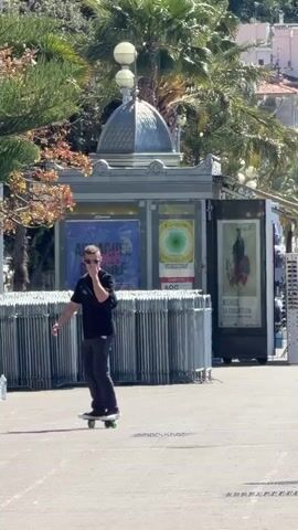The video shows a man riding a skateboard on a paved surface in an outdoor setting. The man is wearing a black t-shirt, gray pants, and sunglasses. He also has a black backpack. He is balancing on the skateboard and using his arms to maintain his balance. There are several people seated on benches in the background, and people are walking past in the foreground. There are also metal barriers and a small building with posters on it. The environment appears to be a public plaza with trees and palm trees nearby. The weather seems sunny and clear.