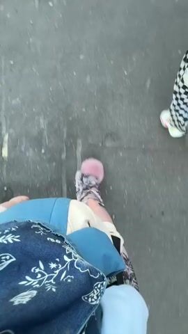 The video is a first-person perspective of someone walking on a paved surface, likely a sidewalk or street. The camera is pointed downwards, focusing on the legs and feet of the person filming, as well as the legs and feet of at least two other individuals walking ahead. The person filming is wearing patterned pants and a jacket with blue and white fabric. The other individuals are wearing pants with a black and white checkerboard pattern and white shoes. The ground is grey and appears to be asphalt. The scene is outdoors.