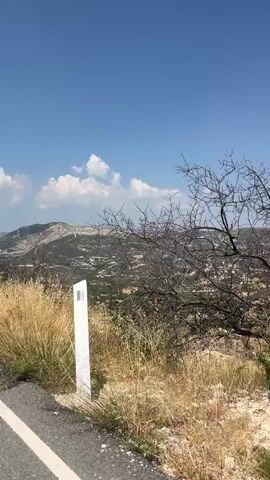 The video is a first-person view from a moving vehicle traveling on a paved road with a white line marking the edge. The landscape outside is a dry, hilly terrain with scattered buildings and vegetation, appearing to be under a clear blue sky. In the foreground, tall, dry grass and some sparse branches are visible. The camera pans slightly, revealing more of the hillside dotted with structures and what appears to be agricultural terraces on a distant hill. The overall impression is of a sunny day in a rural or semi-rural mountainous region.