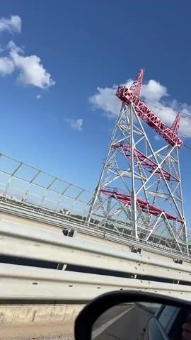The video captures a view from a car driving on a bridge, likely a highway. The primary subject is a large, intricate metal pylon structure, painted white with striking pink accents on its diagonal beams and the upper crossbars. The pylon is immense, reaching high into a bright blue sky dotted with fluffy white clouds, suggesting a clear, sunny day, likely daytime given the lighting. The structure is complex, with multiple levels and angled supports.

The car is on the bridge, indicated by the guardrails and the view of other vehicles passing by. In the foreground, a car's side mirror is visible, reflecting the road and the sky, confirming the perspective. The bridge itself appears to be quite wide, with a fence lining its edge.

There are no visible people in the frame, other than a person in the driver's seat of the car in the left lane, barely visible through the tinted window. The scene evokes a sense of travel and possibly awe at the scale of the industrial structure. The bright colors of the pylon against the natural blue sky create a visually interesting contrast. The location is identified as Novocheboksarsk, Russia, which provides context for the type of infrastructure that might be present.