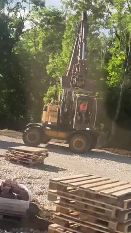 A yellow excavator with a long boom arm is parked on a gravel surface next to a wooded area. The excavator has a cab with an orange-vested operator visible inside. A pile of dirt is located to the right of the excavator. In the foreground, there are stacks of wooden pallets. The scene appears to be a construction or landscaping site.