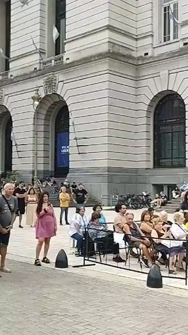 Here is a description of the video:

The video shows an outdoor performance in front of a building with a crowd of people seated and standing. The building has a stone facade and two large arched doors. People are sitting on chairs in front of the building, and some are sitting on the steps. A stage has been set up in front of the crowd. It's built into the back of a truck displaying the emblem of "Secretaria de Cultura." On the stage, two musicians perform. One is playing a trumpet and the other is playing a keyboard. A drum set is also on stage. The sky is overcast.