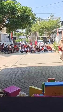 The video shows a street scene with a group of children sitting on the ground, facing a group of adults. The children are dressed in red and white, and some of them are wearing headbands. The adults are also dressed in red and white, and some of them are holding flags. There are buildings and trees in the background. The scene appears to be some kind of celebration or event. In the foreground, there are some colorful boxes and a microphone, suggesting that there might be a performance or presentation taking place. The overall atmosphere seems festive and communal.