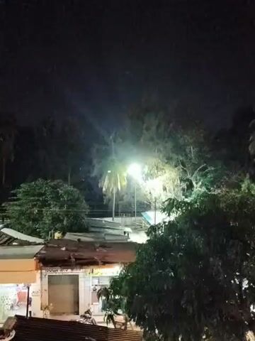 The video shows a nighttime view of a residential or commercial area. A bright street light illuminates a palm tree and surrounding foliage. Below the trees, several buildings with corrugated metal roofs are visible. The buildings appear to be shops or small residences, with storefronts and closed shutters. One building has a yellow facade. The scene is dark, with the street light being the primary source of illumination. The overall impression is of a quiet, possibly rural or semi-urban setting at night.