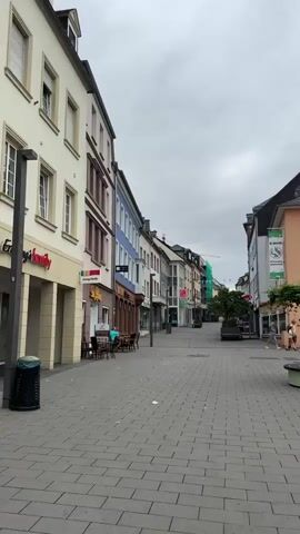 The video pans across a European town square. In the foreground, a person is walking away from the camera, facing down a cobblestone street lined with buildings. The street appears to be a pedestrian zone with shops and businesses. Further into the square, there are outdoor seating areas for cafes and restaurants, some with umbrellas. A food trailer with "Bitburger" branding is visible. The architecture is typical of European towns, with multi-story buildings and a mix of modern and older styles. The sky is overcast. There are no apparent interactions or significant actions taking place. The overall impression is of a quiet, everyday scene in a town.