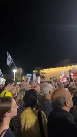 The video shows a crowd of people at night holding signs. An Israeli flag is visible on the left. Some signs have a picture of a man on them with Hebrew text. Another sign says “With NO RELEASE On Sht bites on fire.” There is a palm tree behind the signs and people. The people appear to be looking in the same direction.