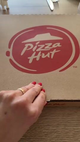 A hand with red nail polish opens a Pizza Hut pizza box on a light brown wooden table. The pizza inside is a large round pizza with a thick crust and a variety of toppings, divided into two different halves. A white plastic pizza saver is placed in the center. The pizza box is then closed. A fork and knife are visible in the background, as is a small white figurine.