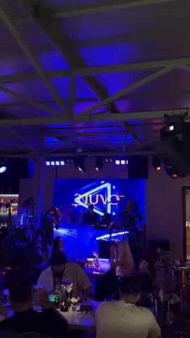 The video shows an indoor venue with a stage and a large LED screen displaying the word "NUVO". A band is performing on stage, with musicians visible behind instruments. The stage is illuminated with blue and purple lights, and there is a haze effect. In the foreground, several people are seated at tables, appearing to be patrons. The overall atmosphere suggests a nightclub or bar setting.
