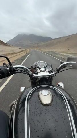 The video is filmed from the perspective of a person riding a black motorcycle with white stripes on a paved road. The rider's hands are visible on the handlebars. In the foreground, the motorcycle's fuel tank, handlebars, and instrument cluster are visible. The road ahead curves to the left and is lined with a white stripe on the left and a dirt shoulder on the right. In the background, there are dry, barren mountains with some vegetation. The sky is overcast with clouds. There appears to be some construction or activity happening in the distance on the right side of the road. The road surface shows signs of being wet, indicating it may have rained recently.