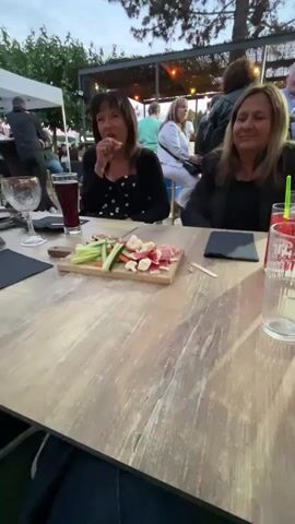 Two women are sitting at a table outdoors, with a platter of food and drinks in front of them. They appear to be in conversation. The woman on the left is wearing a black top with white polka dots, and the woman on the right is wearing a black jacket. In the background, other people are visible, as well as tents and trees. The time of day appears to be late afternoon or early evening, with soft lighting.