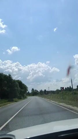 A first-person perspective video shows a car driving on a paved road. The road curves to the left, with a white line marking the lane. On the left side of the road, there are trees and foliage. On the right side, there is a ditch with tall grass and trees further in the distance. There are also power poles and road signs visible on the right. The sky is blue with scattered white clouds, indicating a sunny day. A blurred reddish object is visible in the upper right quadrant of the frame, possibly a smudge or artifact on the windshield.