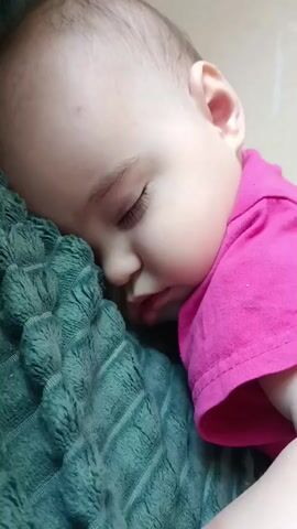 A close-up shot focuses on a sleeping infant. The baby's face is resting on a textured, teal-colored blanket. The infant is wearing a bright pink t-shirt. The image is a still frame from a video, as indicated by the timestamp in the bottom left corner.
