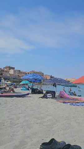 The video shows a crowded beach with people sunbathing, swimming, and relaxing under umbrellas. There are several beach umbrellas, towels, and beach bags scattered across the sand. The water is blue and clear, with some boats visible in the distance. The sky is clear and blue, with a few scattered clouds. People are dressed in swimwear, and some are playing in the water. There is a ferry with smoke coming out of its stack in the distance. The overall atmosphere is one of leisure and enjoyment on a sunny day at the beach.