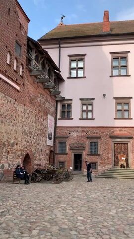 The video shows the courtyard of a castle. The courtyard is paved with cobblestones. The castle buildings are made of red brick and painted in light pink and white. There are several people walking around the courtyard. Two people are sitting on a bench next to an old-fashioned horse-drawn carriage. A man is standing in front of the entrance to a building. A couple is walking hand in hand. A man in sunglasses is walking by himself. The sky is blue.