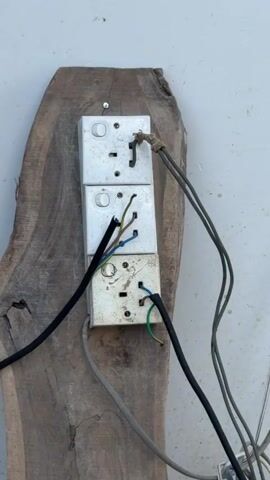 A stacked electrical outlet assembly is mounted on a piece of wood that is attached to a wall. The outlets are dirty and appear to be in disrepair. Multiple electrical wires are connected to the outlets and are running in various directions. A man is standing in front of the outlets, facing away from the camera, with his hands behind his back. He is wearing a dark blue shirt and dark blue pants. The environment appears to be an outdoor construction site, with debris and building materials visible in the background.
