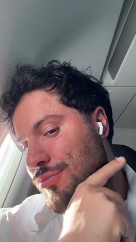 A close-up video shows a man with short dark hair and a beard wearing a white collared shirt, sitting on an airplane. He is wearing a white earbud in his right ear. He looks to the side, then closes his eyes briefly. He then looks into the camera, closes his eyes again, and puckers his lips. Sunlight shines through the airplane window.