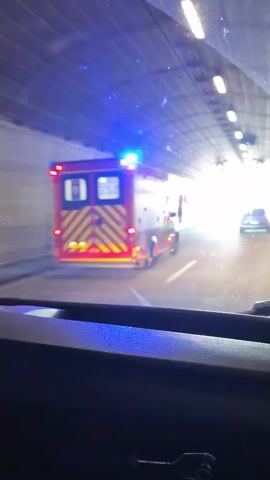 The video shows a car driving through a tunnel and then onto a bridge. The bridge is supported by tall concrete pillars with metal cable stays.  An ambulance with flashing blue lights is ahead of the car. There are other cars on the bridge with the car. The weather appears to be sunny.