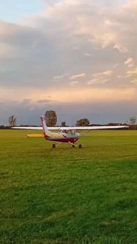 A small red and white airplane is parked on a grassy field. The sky is cloudy and appears to be either sunrise or sunset. The plane is facing forward and appears to be stationary. There are trees and a fence in the background. The grass is green and appears to be well-maintained. The overall scene is peaceful and serene.