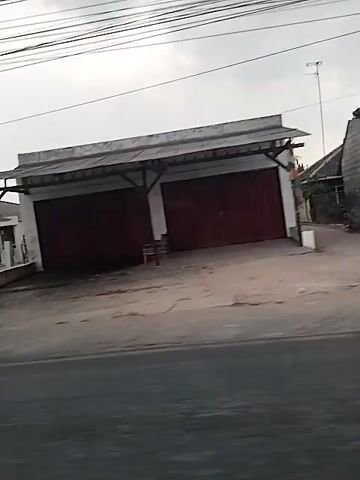 The video shows a roadside view of a street with a few buildings and vegetation. On the left side, there is a small structure with a canopy, possibly a street food stall. Further down the road, there are several buildings with closed doors and windows, suggesting they might be shops or commercial establishments. In the background, there are trees and a cloudy sky. The camera appears to be moving, giving a sense of passing by these locations. The overall scene depicts a quiet, possibly rural or suburban, street.
