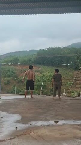 A man and a woman stand on a concrete surface with puddles of water. The man, shirtless and wearing black shorts, faces away from the camera. The woman, wearing a brown jumpsuit, also faces away from the camera. They appear to be performing calisthenics or a similar physical activity. In the background, there is a lush green hillside with trees and some structures. The sky is overcast and cloudy. The scene suggests an outdoor environment, possibly in a rural or suburban setting.