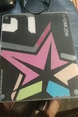 The video is an extreme close-up, slightly shaky, top-down view of a G-Star 2016 limited edition item. It appears to be a laptop case or sticker, featuring a large, multi-colored star design in pink, beige, light green, and light blue, outlined in black and grey. The background is black with white angular shapes. There are visible signs of wear and tear on the item, with peeling paint or stickers in several areas. The edge of a dark object, possibly a computer mouse, is visible in the top left corner, and a black cable extends from it. A portion of a person's foot and toes are visible in the bottom center of the frame, indicating the item is on a surface where someone is present. The lighting is dim, creating shadows.