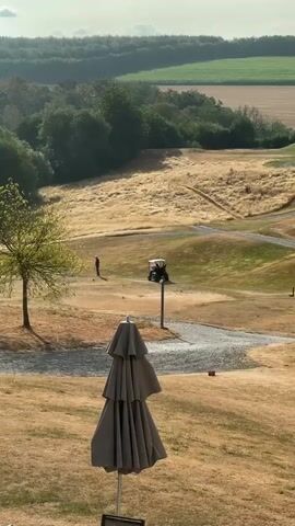A video shows a golf course on a sunny day. In the foreground, a closed umbrella is in focus, obscuring some of the view. In the middle ground, a single person is standing on a golf tee box, appearing to be about to hit a golf ball. A golf cart is parked nearby on a path. The landscape of the golf course consists of rolling hills with dry, golden grass and some patches of green. Trees are visible on the left side of the frame, and a distant treeline marks the horizon. The overall impression is a calm, outdoor recreational setting.
