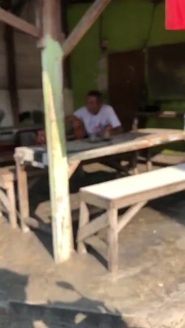 A slightly blurry video shows an outdoor seating area of what appears to be a small food stall or eatery. In the background, a man wearing a white t-shirt is seated at a table, looking down at something in front of him. He is the only person clearly visible. The table is covered with a light-colored tablecloth or surface. To the right of the table, a wooden bench is present. The environment appears rustic, with wooden beams and posts supporting a roof. The ground is concrete. Sunlight casts shadows, indicating it is daytime. The overall impression is a simple, casual setting.