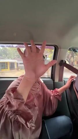 A person in a pink hijab and dress sits in the back seat of a vehicle, holding their hand up to cover their face. They are looking out the window. Another person, wearing a dark burgundy hijab, is visible behind them, leaning forward. The interior of the car is dimly lit, with visible seatbelts and headrests.