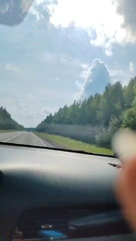 The video shows a view from inside a car, looking out the windshield. The car appears to be driving on a highway through a wooded area. The sky is blue with some fluffy white clouds, and the sun is shining, creating lens flare in the footage. The trees on the side of the road are lush and green. The dashboard of the car is visible in the foreground. The perspective suggests the footage was captured by a passenger or a dashcam.