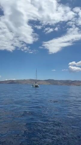 A white catamaran sails across a calm blue sea under a bright, partly cloudy sky. The vessel is moving away from the viewer towards a distant coastline characterized by rugged, light-brown hills and mountains. The sky is predominantly clear blue with scattered white, cumulus clouds. The water exhibits gentle ripples, reflecting the clear sky. The scene depicts a serene daytime setting in Koundouros, Greece. There is no visible text.