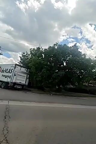 The video is shot from a moving vehicle looking out a side window. The video begins by showing the left side of the road, with a sidewalk and some trees. It then pans to the right, showing more trees and a grassy area. The sky is cloudy with some patches of blue. There are a few people on the side of the road in the beginning of the video, one person on a scooter or bike and another person who appears to be sitting or crouching. The video continues to pan to the right, showing more of the treeline and grass. The movement of the vehicle obscures some of the details in the video, but it appears to be a rural or suburban setting.