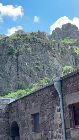 The video captures an outdoor scene at Geghard Monastery. The footage begins by showcasing the monastery's stone walls and a tall, steep cliff behind it, with a white cross visible on the cliff face. A metallic downspout is also seen on the building. The camera then pans right, revealing the lush green mountainside and several people, including a person recording with a phone. The camera continues to pan, showing more of the monastery's architecture, including an ornate arched entrance and another building with scaffolding. More people are visible around the entrance. The video then pans down a path with a stone wall and some trees, showing more people walking and relaxing. The camera then pans up to show more of the majestic rock formations and vegetation. Finally, the camera zooms out, revealing a large stone archway and more of the surrounding natural landscape. The sky is bright blue with scattered white clouds throughout the video.