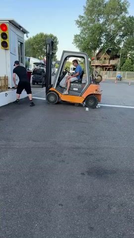 A man in a blue t-shirt and white shorts operates an orange and grey forklift. He is sitting in the driver's seat, holding the steering wheel. Beside him, a second man in a black t-shirt and black shorts stands near the side of the forklift. The forklift is positioned on an asphalt surface, with white painted lines indicating parking spaces or a driving lane.

In the background, a small white booth or building is visible, featuring a traffic light with a red light illuminated. A small wooden fence runs along the front of the booth. Further back, trees and a wooden structure resembling a treehouse are visible, suggesting an outdoor recreational area. A large red planter with some greenery is also present.

The forklift appears to be moving towards or away from the white booth. The overall scene suggests an operational or setup activity at an outdoor venue. The lighting indicates it is daytime, likely late afternoon or early evening, with a clear sky. The setting could be a fairground, amusement park, or similar event space. No discernible emotions are evident from the individuals, but their focus seems to be on the task at hand.

Visible text on the forklift reads "STILL R70-35T".