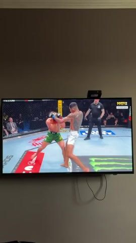 A television mounted on a wall shows a UFC match. One fighter is wearing green shorts and the other white shorts. The fighter in white shorts throws a kick toward the other fighter's head. A referee stands in the ring. The audience is visible beyond the ring. Text is visible on the screen, including "UFC FIGHT NIGHT," "МАТЧ!" and "PMARKETS.COM." Cables are visible hanging below the television. A black device is mounted on top of the television. The wall is a light neutral color.