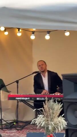 A man in a black suit jacket over a white shirt is seated at a red Nord Stage 4 keyboard, playing and singing into a microphone. He is on a stage with a beige backdrop and a string of lights above. A large speaker is visible to the right, and a bouquet of dried flowers is in front of the keyboard.