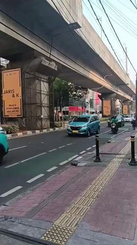 The video shows a street scene with an elevated structure overhead, likely a highway or train line. Several vehicles, including cars and motorcycles, are moving on the road. A blue car is in the foreground, followed by other cars and motorcycles. On the sidewalk, there are tactile paving blocks, indicating a pedestrian area, and bollards are present. Advertisements are visible on the pillars supporting the elevated structure. The overall environment appears to be urban.