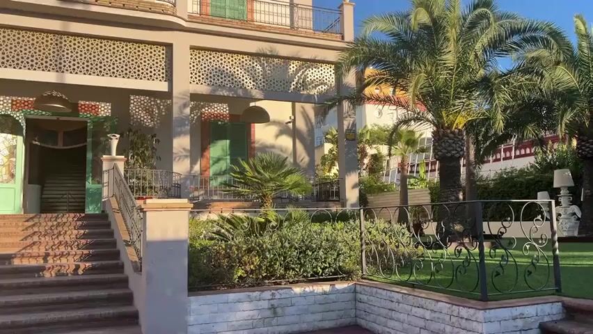 The video pans across the exterior of a multi-story building with a distinctive architectural style. The building has a light-colored facade with ornate, patterned screens over some of the windows and balconies. Green shutters are visible on some windows, and a balcony with a black railing extends across the upper level. In front of the building, a set of brick stairs leads up to an entrance with a light green door. A small garden with various plants and palm trees is situated to the right of the stairs. To the left of the main building, a red building with similar architectural elements, including green shutters and a balcony, is visible. The scene is bathed in sunlight, suggesting it is daytime. The sky is clear and blue. The ground in front of the red building is a red-toned pathway. The overall impression is of a well-maintained, possibly residential or hospitality property in a warm climate.