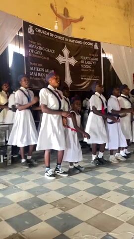 A group of about ten children, mostly girls, are singing and clapping in a church. They are wearing white dresses or skirts with dark accents like belts and ties. One girl is wearing a bright red tie. Some of them are wearing dark shoes and others are wearing sneakers. They are standing on a checkered floor in front of a banner that reads "Church of Christ in Nations (COCIN)" and has a large cross in the center. There's a figure of Jesus with outstretched arms depicted above the banner. The church has a simple interior with white drapes.