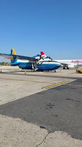 A large blue and white amphibious aircraft, a Bombardier CL-415, is parked on an airport tarmac under a clear blue sky. The aircraft has its door open, and a set of stairs leads up to it. A passenger jet with "Austrian" written on its side is visible behind the amphibious aircraft. The tarmac is marked with yellow and grey lines. The scene appears to be a sunny day at an airport.