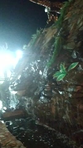 The video shows a close-up view of water trickling down a rocky, textured surface that appears to be part of a decorative element, possibly in an indoor environment or a simulated natural setting. Some green artificial leaves are interspersed among the rocks and the water. The lighting suggests it might be nighttime or a dimly lit indoor space, with a bright light source visible in the background, creating a lens flare effect. The overall impression is one of a water feature or a decorative rock formation with water flowing over it. There are no people or animals visible in the video.