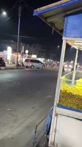 The image captures a nighttime street scene in Gampongjawa, Indonesia, from the perspective of someone standing behind a street vendor's cart. The cart, predominantly blue and white, features an illuminated display case filled with what appear to be yellow, shelled peanuts or similar snacks.

The street is a paved road, busy with vehicular traffic. Numerous motorcycles, many carrying multiple riders, are seen moving in both directions, their headlights and taillights creating streaks of light. Cars, including a silver SUV and a darker sedan, are also present, navigating the road. The general lighting is provided by streetlights, with a prominent tall, multi-armed streetlight visible on the left, and illuminated storefronts of buildings lining both sides of the street in the background. These buildings suggest a commercial or residential area. Overhead, power lines crisscross the dark sky. The road appears dry, indicating clear weather. No specific activities other than routine traffic flow are discernible. There is no clearly legible text visible in the frame.