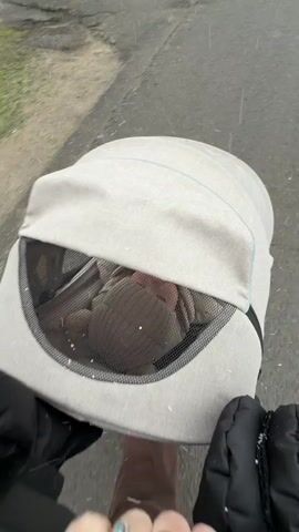 A first-person point-of-view video shows an individual pushing a baby in a light-colored stroller along an outdoor path or road in Reshetnikovo, Russia. The person's hands, wearing black gloves, are visible on the stroller handle. A gold ring with a blue stone is on the left hand, and a plain gold band is on the right.

Light snow or sleet is falling, with small white flakes visible on the stroller's canopy and falling into the mesh viewing window. Through this window, a baby's head is visible, wearing a dark-colored hat or hood. The baby appears to be resting or sleeping calmly, with its face partially obscured but hinting at a peaceful expression. The ground beneath the stroller is dark and appears wet, possibly from the precipitation. The overall scene depicts a quiet, wintry walk.