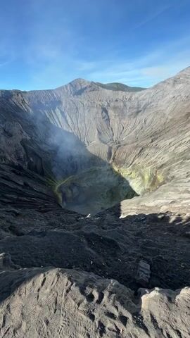 This video shows a view from the rim of a volcanic crater. The crater is a large, circular depression with steep, barren walls made of dark rock and ash. The bottom of the crater is not fully visible, but it appears to be filled with a greenish-yellow substance, possibly sulfur deposits, and there is a plume of white smoke rising from its center. The sky above is a clear, bright blue with a few wispy clouds. The foreground of the image shows a sandy, uneven surface, possibly the rim of the crater, with tire tracks or footprints visible in the sand. The lighting suggests it is daytime, with the sun illuminating the crater from the side, casting shadows on the walls. The overall impression is one of a rugged, active, and awe-inspiring natural landscape.