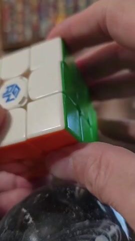 A person's hands are manipulating a white, orange, and green Rubik's Cube, with a blue logo visible on one of the white faces. The background is out of focus but suggests a home environment with shelves of items. The cube is being turned and twisted.