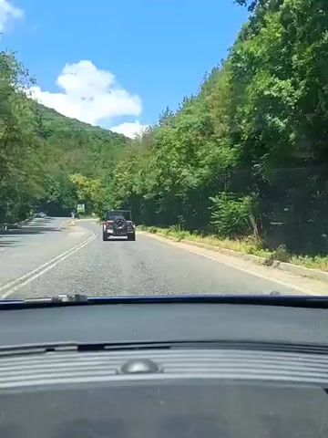 A video taken from the perspective of a vehicle's dashboard shows a paved road winding through a lush, green, mountainous area. The sky is blue with scattered white clouds. The vehicle is following a dark colored Jeep Wrangler. There are trees and vegetation on both sides of the road, with the left side appearing to be a steep hillside. Traffic signs are visible on the sides of the road. The video appears to be from a sunny day.
